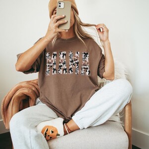 May include: A brown t-shirt with a white and black graphic print that says "Mama" with a Halloween theme. The graphic print features pumpkins, bats, and ghosts.
