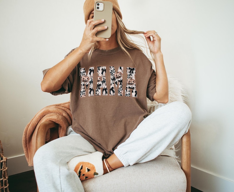 May include: A brown t-shirt with a white and black graphic print that says "Mama" with a Halloween theme. The graphic print features pumpkins, bats, and ghosts.