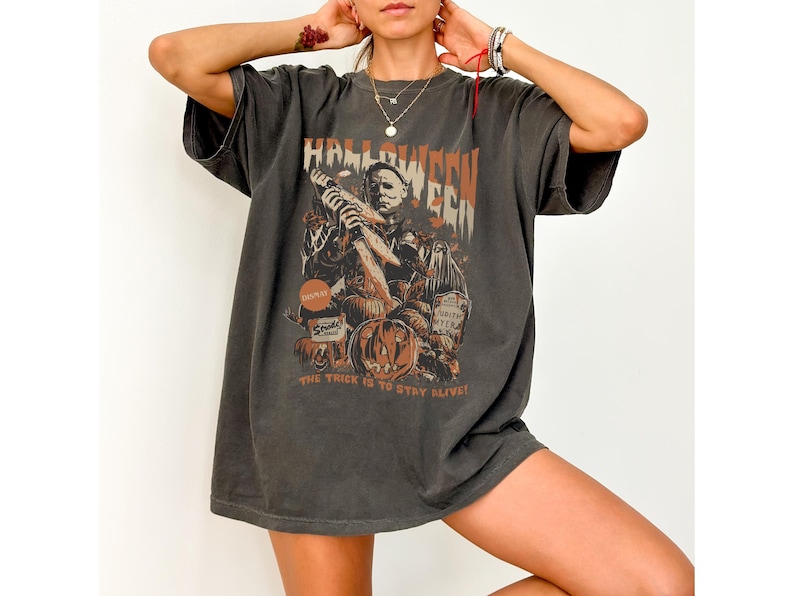 Puede incluir: Una camiseta gris oscuro de gran tama&ntilde;o con un estampado gr&aacute;fico de un personaje de pel&iacute;cula de terror que sostiene un cuchillo. El texto "Halloween" est&aacute; impreso sobre el personaje y "The trick is to stay alive!" est&aacute; impreso debajo del personaje.