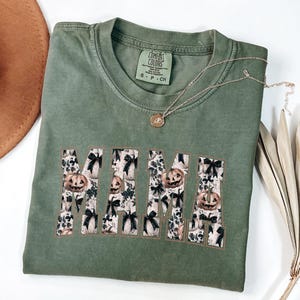 May include: Green t-shirt with the word "MAMA" in large, white letters. The letters are filled with a pattern of black bows and orange pumpkins. The shirt is made by Comfort Colors.