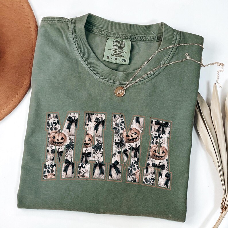 May include: Green t-shirt with the word "MAMA" in large, white letters. The letters are filled with a pattern of black bows and orange pumpkins. The shirt is made by Comfort Colors.