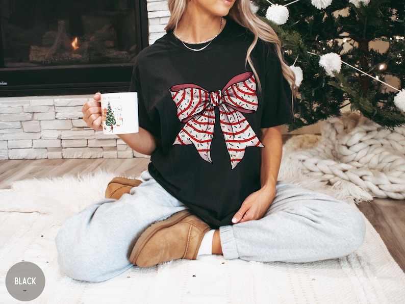 May include: A black t-shirt with a red and white bow with a sprinkle pattern. The person wearing the shirt is sitting on a white fuzzy rug.