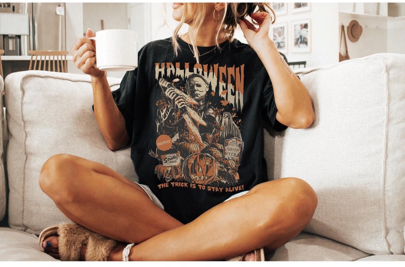 Puede incluir: Una camiseta negra con un estampado gr&aacute;fico de un personaje de pel&iacute;cula de terror sosteniendo un cuchillo. El texto "Halloween" est&aacute; impreso sobre el personaje y el texto "The trick is to stay alive!" est&aacute; impreso debajo del personaje.