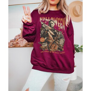 May include: Burgundy Halloween sweatshirt with a graphic of a horror movie character holding a knife, a carved pumpkin, and the text "Halloween". The text "The trick is to stay alive!" is also printed on the sweatshirt.