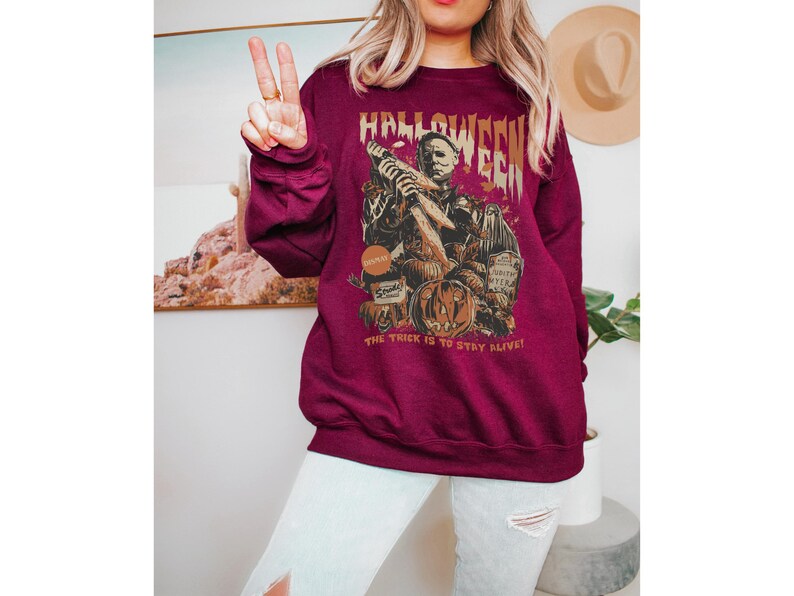 May include: Burgundy Halloween sweatshirt with a graphic of a horror movie character holding a knife, a carved pumpkin, and the text "Halloween". The text "The trick is to stay alive!" is also printed on the sweatshirt.