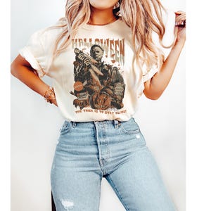 Puede incluir: Una camiseta blanca con un estampado gr&aacute;fico de un personaje de pel&iacute;cula de terror sosteniendo un arma. El texto "Halloween" est&aacute; impreso sobre el personaje y "The trick is to stay alive!" est&aacute; impreso debajo del personaje.