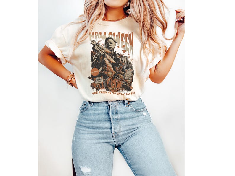 Puede incluir: Una camiseta blanca con un estampado gr&aacute;fico de un personaje de pel&iacute;cula de terror sosteniendo un arma. El texto "Halloween" est&aacute; impreso sobre el personaje y "The trick is to stay alive!" est&aacute; impreso debajo del personaje.