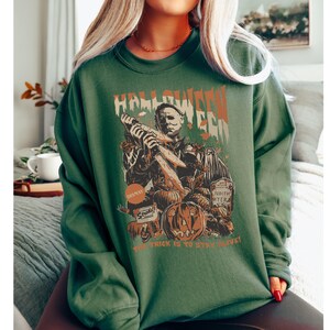 May include: Green long-sleeve sweatshirt with a Halloween-themed graphic. The design features a depiction of Michael Myers holding a knife, pumpkins, and the text "Halloween Era." The phrase "The trick is to stay alive!" is also included.