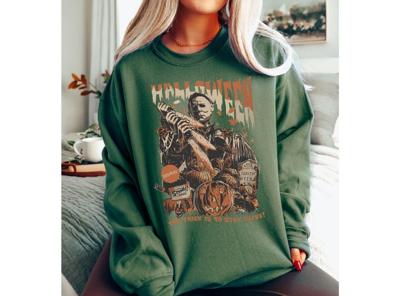 May include: Green long-sleeve sweatshirt with a Halloween-themed graphic. The design features a depiction of Michael Myers holding a knife, pumpkins, and the text "Halloween Era." The phrase "The trick is to stay alive!" is also included.