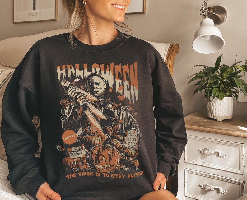 May include: Black Halloween sweatshirt featuring a graphic of Michael Myers holding a knife, pumpkins, and a tombstone. The word "Halloween" is printed across the top in orange and white. The phrase "The trick is to stay alive!" is printed at the bottom.