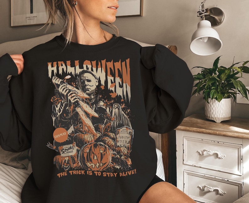 May include: Black Halloween-themed sweatshirt with a graphic featuring a menacing figure holding knives, pumpkins, and the text "HALLOWEEN". The phrase "THE TRICK IS TO STAY ALIVE!" is also printed on the sweatshirt.