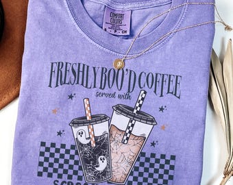 May include: A lavender Comfort Colors t-shirt with the text "FRESHLY BOO'D COFFEE served with SCREAM & SUGAR." The graphic features two iced coffee drinks with ghost and spiderweb designs. A gold necklace rests on the shirt.