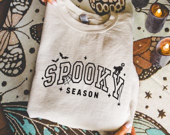 May include: A cream-colored sweatshirt with the words "SPOOKY SEASON" in black block letters. The design includes bats, a spiderweb, a black cat, and stars. The sweatshirt is folded and resting on a patterned surface.