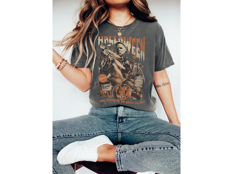 Puede incluir: Una camiseta gris con un gr&aacute;fico vintage de Halloween que presenta una figura enmascarada sosteniendo un cuchillo. El texto en la camiseta dice "Halloween" y "The trick is to stay alive".