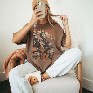 Puede incluir: Una mujer lleva una camiseta marr&oacute;n con un gr&aacute;fico de un personaje de la pel&iacute;cula Halloween. La camiseta dice "Halloween". Lleva pantalones de ch&aacute;ndal blancos y un par de calcetines con tem&aacute;tica de calabaza.