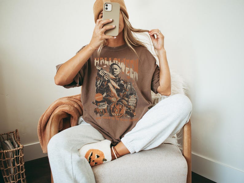 Puede incluir: Una mujer lleva una camiseta marr&oacute;n con un gr&aacute;fico de un personaje de la pel&iacute;cula Halloween. La camiseta dice "Halloween". Lleva pantalones de ch&aacute;ndal blancos y un par de calcetines con tem&aacute;tica de calabaza.