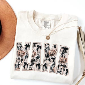 May include: A cream colored t-shirt with the word "MAMA" printed in black. The letters are filled with a pattern of black bows and orange pumpkins. The shirt has a small tag that says "COMFORT COLORS" on it.