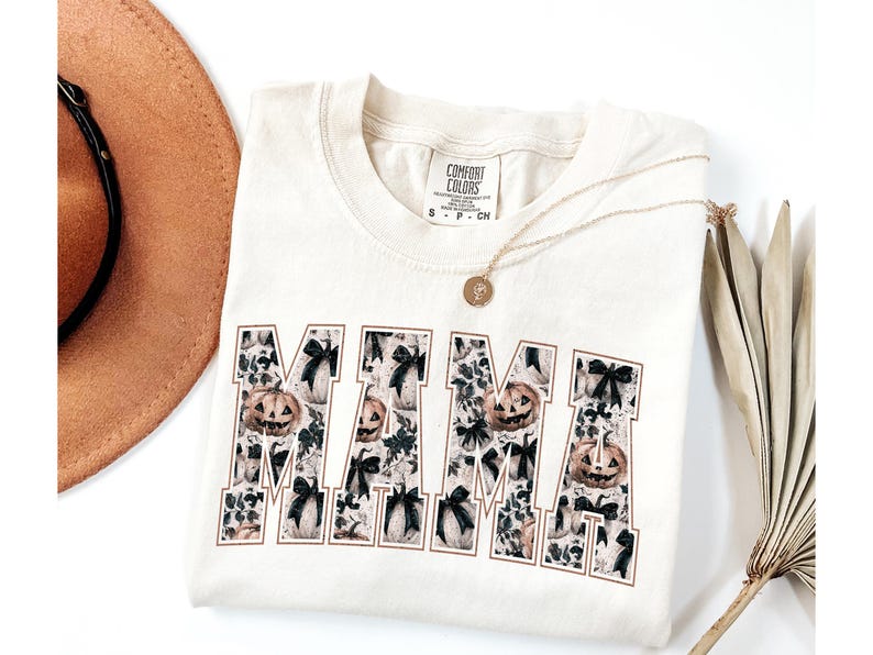 May include: A cream colored t-shirt with the word "MAMA" printed in black. The letters are filled with a pattern of black bows and orange pumpkins. The shirt has a small tag that says "COMFORT COLORS" on it.