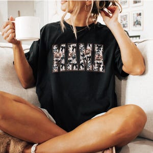 May include: A black t-shirt with a graphic print of the word "MAMA" in white letters with a Halloween theme. The letters are decorated with pumpkins, bats, and other Halloween imagery.