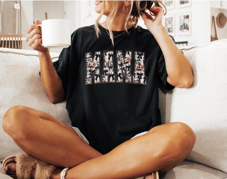 May include: A black t-shirt with a graphic print of the word "MAMA" in white letters with a Halloween theme. The letters are decorated with pumpkins, bats, and other Halloween imagery.