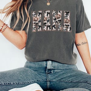 May include: A dark gray t-shirt with a graphic print of the word "MAMA" in white with a black outline. The letters are filled with a pattern of pumpkins and bows.