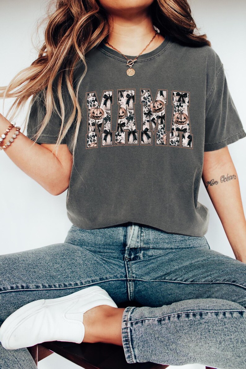 May include: A dark gray t-shirt with a graphic print of the word "MAMA" in white with a black outline. The letters are filled with a pattern of pumpkins and bows.