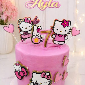 May include: A pink cake decorated with Hello Kitty-themed toppers and accents. The cake features a gold number 7, the name "Ayla", and several Hello Kitty figures. Pink hearts and Hello Kitty images adorn the sides.