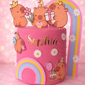 Può includere: Una torta di compleanno rosa decorata con personaggi di orsi dei cartoni animati, arcobaleni e margherite. La torta ha il nome "Sophia" in oro. Gli orsi tengono in mano regali, fiori e un cupcake. Un arco arcobaleno è sul lato.