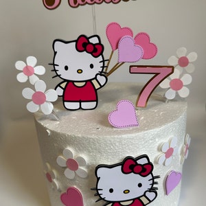 May include: A white cake decorated with Hello Kitty characters, pink hearts, white flowers, and a gold number 7. The cake is topped with a brown and pink sign that says "Helloisa".