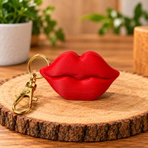 Puede incluir: Un llavero tridimensional en forma de labios de color rojo brillante con un acabado mate suave. Los labios están unidos a una anilla de metal dorado y a un cierre de mosquetón a juego, lo que permite sujetarlo de forma segura a llaves o bolsos.