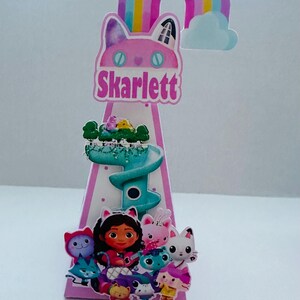 May include: A pink and white cone-shaped party decoration featuring a cartoon cat face with the name "Skarlett" in pink text. The decoration includes a rainbow, a cloud, and various cartoon characters. The overall theme is a children's birthday party.