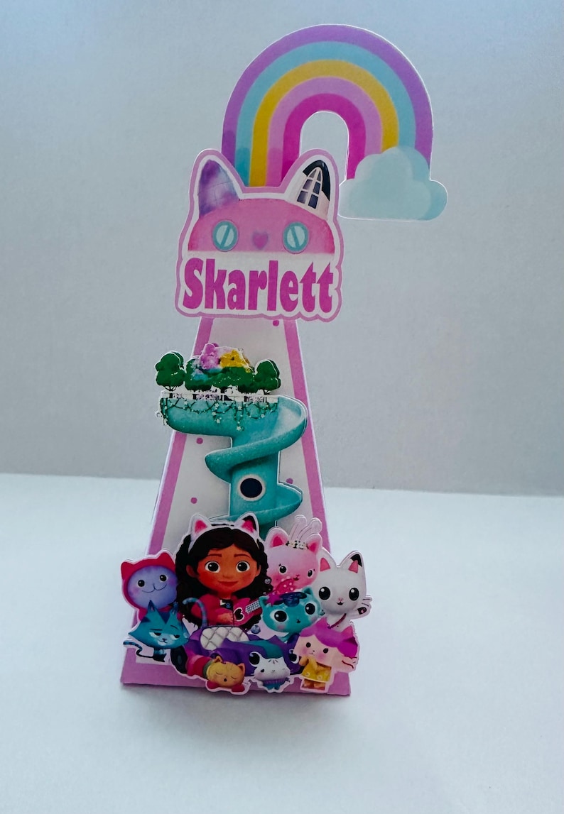 May include: A pink and white cone-shaped party decoration featuring a cartoon cat face with the name "Skarlett" in pink text. The decoration includes a rainbow, a cloud, and various cartoon characters. The overall theme is a children's birthday party.