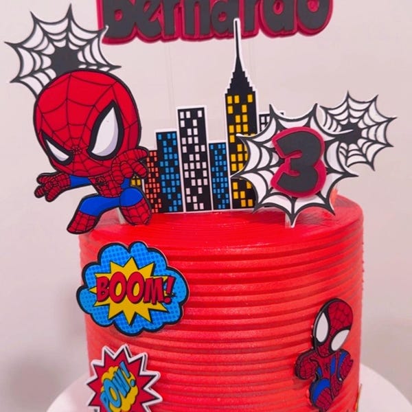 Może przedstawiać: Czerwony tort urodzinowy ozdobiony topperami w stylu Spider-Mana. Ciasto zawiera panoramę miasta, imię "Bernardo" i liczbę "3". Zawiera również grafiki w stylu komiksowym "BOOM!" i "POW!"