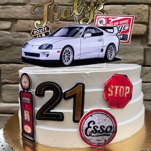 May include: A white frosted cake decorated with a white car, the number "21", and vintage-style signs. The cake has a gold cake board and is decorated with a car, a stop sign, and a gas pump.
