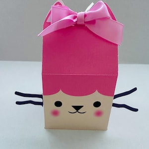 May include: A pink gift box with a cartoon cat face design. The box has a pink bow on top and black whiskers. The cat face features black eyes, a black nose, and pink cheeks. The box is made of paper.