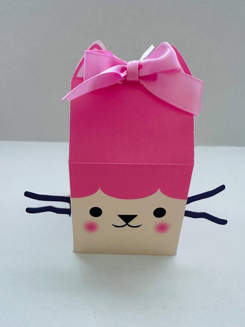 May include: A pink gift box with a cartoon cat face design. The box has a pink bow on top and black whiskers. The cat face features black eyes, a black nose, and pink cheeks. The box is made of paper.