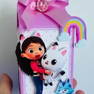 May include: A decorative gift box featuring a cartoon girl hugging a white cat. The box is lavender with a pink bow and a jewel accent. A rainbow and a blue cat are also featured. The box is held by a hand.