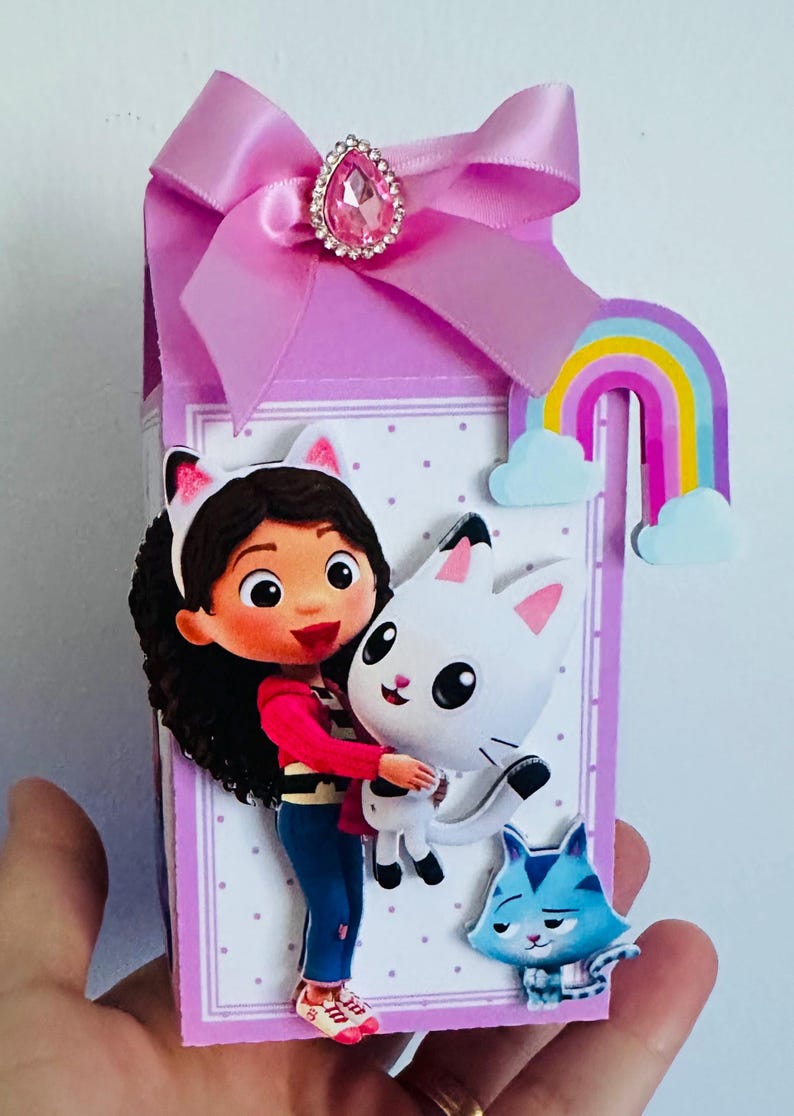 May include: A decorative gift box featuring a cartoon girl hugging a white cat. The box is lavender with a pink bow and a jewel accent. A rainbow and a blue cat are also featured. The box is held by a hand.