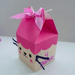 May include: A pink and tan gift box with a cartoon face and a pink bow. The box has a scalloped edge and the number "4" with the name "Skarett" on the side. The box is designed for a birthday or special occasion.