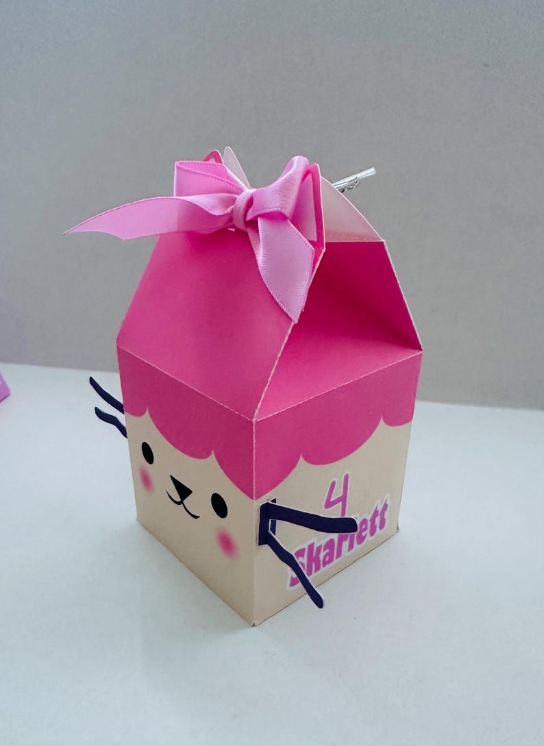 May include: A pink and tan gift box with a cartoon face and a pink bow. The box has a scalloped edge and the number "4" with the name "Skarett" on the side. The box is designed for a birthday or special occasion.