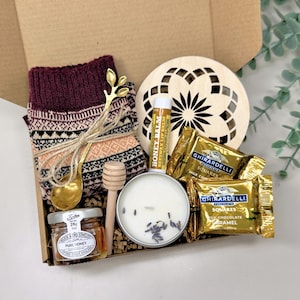 May include: A gift box filled with cozy items. Includes a pair of burgundy patterned socks, a gold-colored spoon, a small jar of honey, a wooden honey dipper, a candle, lip balm, a wooden coaster, and Ghirardelli chocolate squares.
