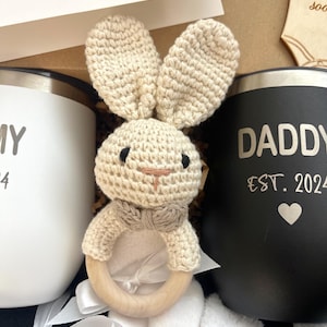 May include: A gift basket for a new parent includes a white mug with the word "Mommy" and a black mug with the words "Daddy Est. 2024" on it. A crocheted bunny rattle with a wooden ring is in the centre of the basket.