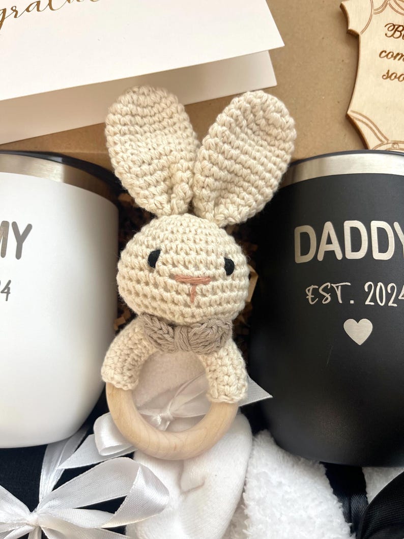 May include: A gift basket for a new parent includes a white mug with the word "Mommy" and a black mug with the words "Daddy Est. 2024" on it. A crocheted bunny rattle with a wooden ring is in the centre of the basket.