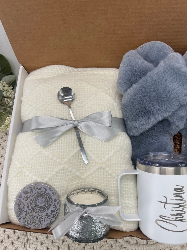 Op de afbeelding: Een cadeaudoos gevuld met knusse items, waaronder een wit gebreid deken, een zilveren kaars, een zilveren mok met de naam "Christina" erop, een zilveren lepel en een paar grijze fluffy pantoffels.