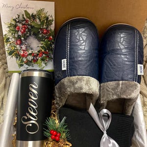 Pode incluir: Uma caixa de presente cheia de uma caneca de viagem preta personalizada com o nome "Steven" gravado nela, um par de pantufas azuis com "HOME" escrito nelas, um par de meias pretas e um cart&atilde;o de Natal com uma coroa.