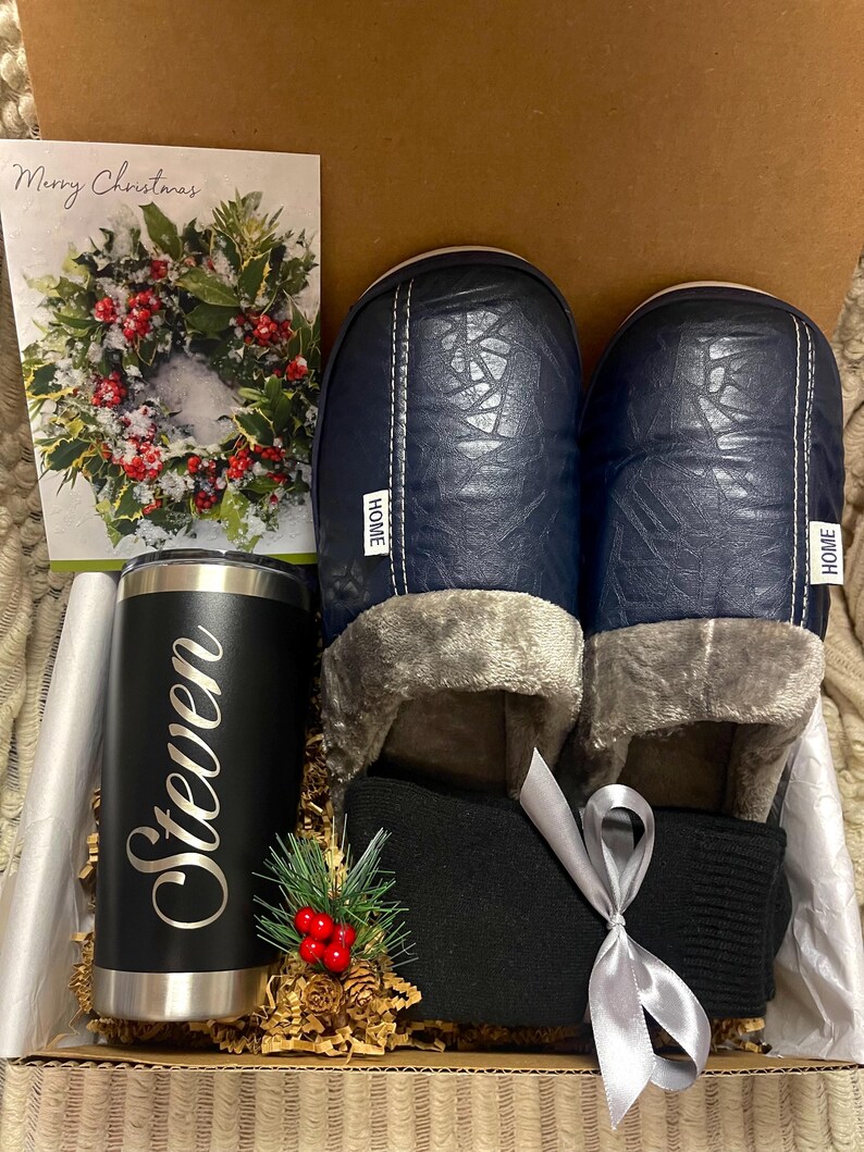 Pode incluir: Uma caixa de presente cheia de uma caneca de viagem preta personalizada com o nome "Steven" gravado nela, um par de pantufas azuis com "HOME" escrito nelas, um par de meias pretas e um cart&atilde;o de Natal com uma coroa.