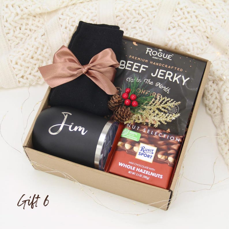Puede incluir: Una caja de regalo que contiene un vaso negro con "Jim" en escritura plateada, calcetines negros con un lazo color rosa, carne seca y una barra de chocolate. La caja est&aacute; decorada con pi&ntilde;as y vegetaci&oacute;n festiva.