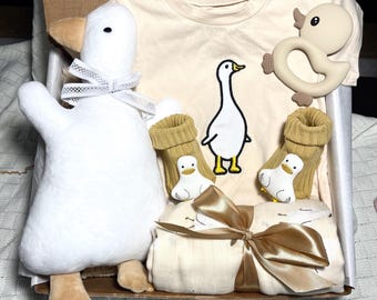 May include: A gift box filled with baby items, including a white plush goose toy, a cream colored t-shirt with a goose design, a pair of yellow booties, and a cream colored blanket with a gold ribbon.