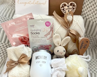 May include: A white gift basket filled with items for a new mom. The basket includes a white tumbler with the text "Mommy Est. 2024", a pair of white socks, a white washcloth, a white crocheted bunny, a wooden spoon with the text "Anderson Baby", a white blanket, and a pink and white hydrating socks package.