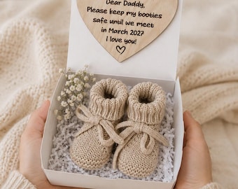 Peut inclure: Une boîte cadeau blanche contenant une paire de chaussons pour bébé en tricot beige avec des lacets, posée sur des lambeaux de papier blanc. Une plaque en bois en forme de cœur est fixée au couvercle, gravée avec: Dear Daddy, Please keep my booties safe until we meet in March 2027 I love you!